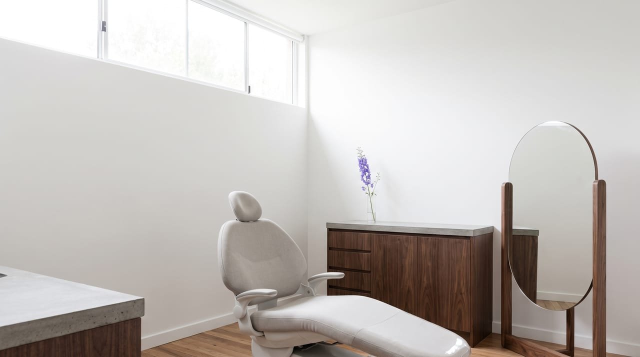 A consultation room at Larkspur: white walls, a tall clerestory window, a dental chair in pale stone upholstery, walnut cabinetry, a large round mirror on a stand and a single stem of larkspur in a glass vase