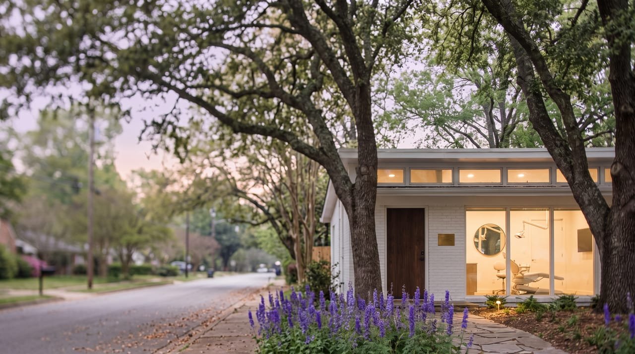 Larkspur Smile Studio at early dusk, warm light in the front window showing the chair, lamp and mirror, larkspur lit by a garden light under the oaks