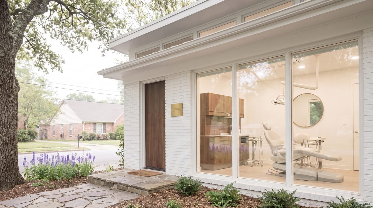The front of Larkspur Smile Studio on a spring morning: a white brick former portrait studio with a walnut door, a bed of violet larkspur, and a wide window showing a pale grey dental chair, an overhead lamp and a tall round mirror in a bright white room