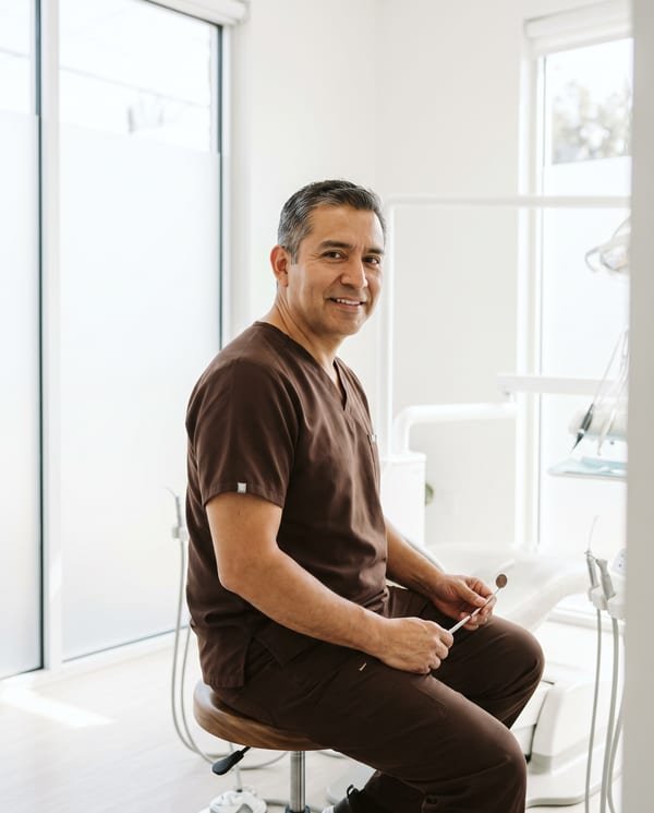 Portrait of Theo Castellanos, DDS, general dentist at Larkspur, seated on a walnut stool in a bright white studio